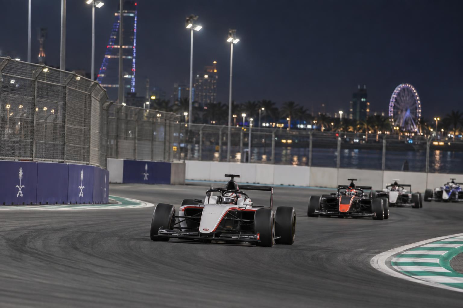 Formula 4 cars race through a floodlit corner at Jeddah Corniche Circuit.
