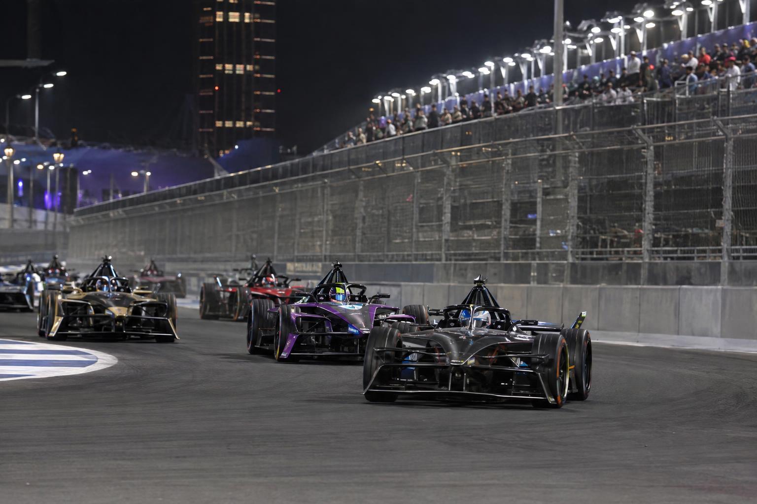 A pack of GEN4 Formula E cars races through a floodlit Jeddah Corniche Circuit corner.